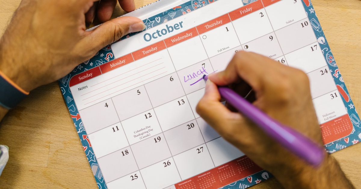Close-up of a person writing a lunch reminder on an October calendar with a purple pen.